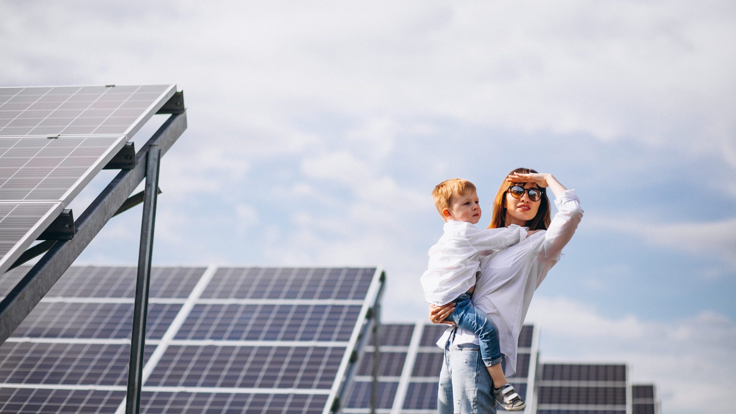 solar-panels-senivpetro Woman holding child with solar panels in the background illustrates blog "How Do Solar Roof Panels Work?"